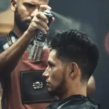 barber using styling spray on young man with black hair, short on sides and long in a quiff on top